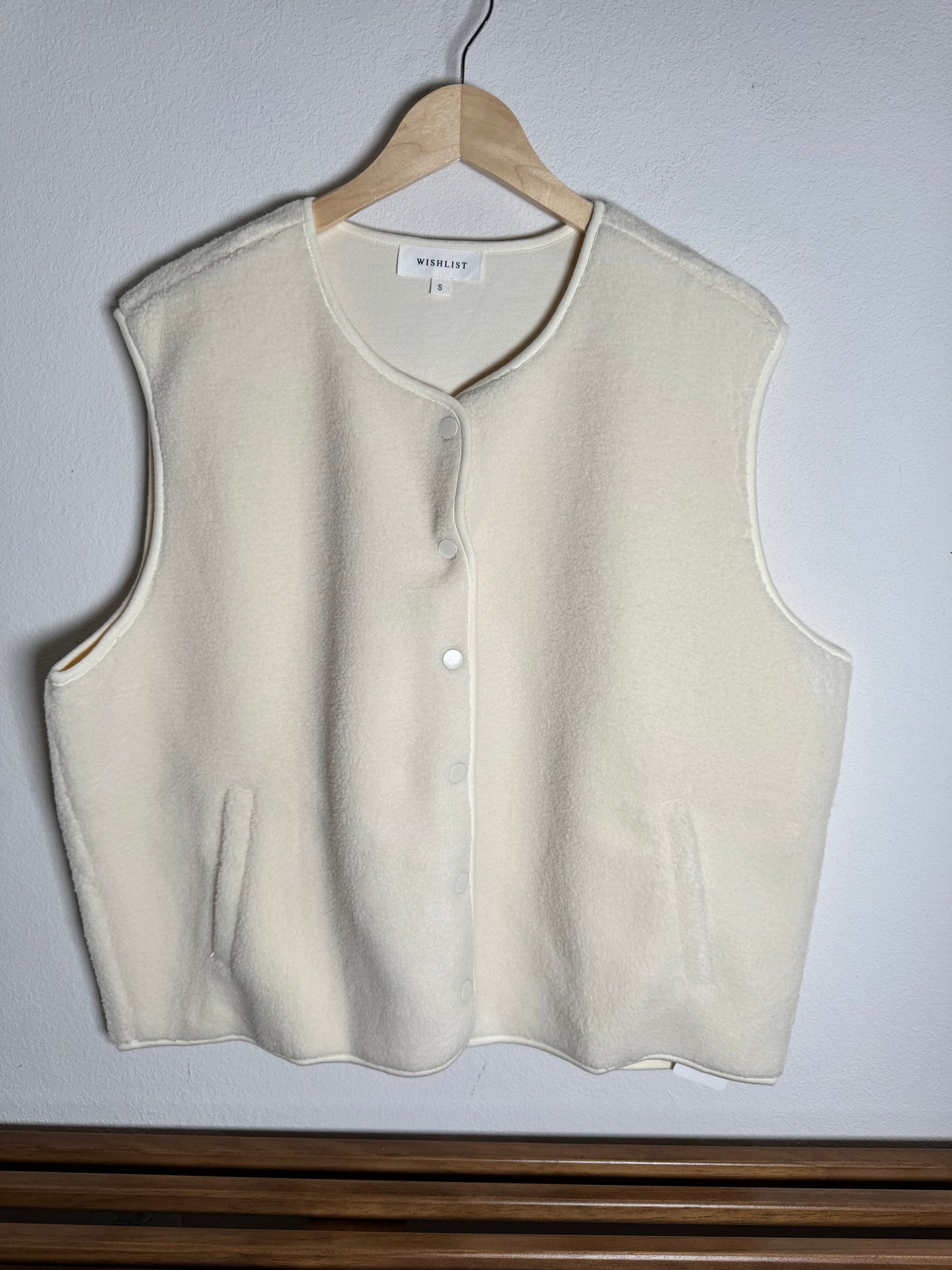 Cream Sherpa sleeveless snap front vest shown in front of a white wall 