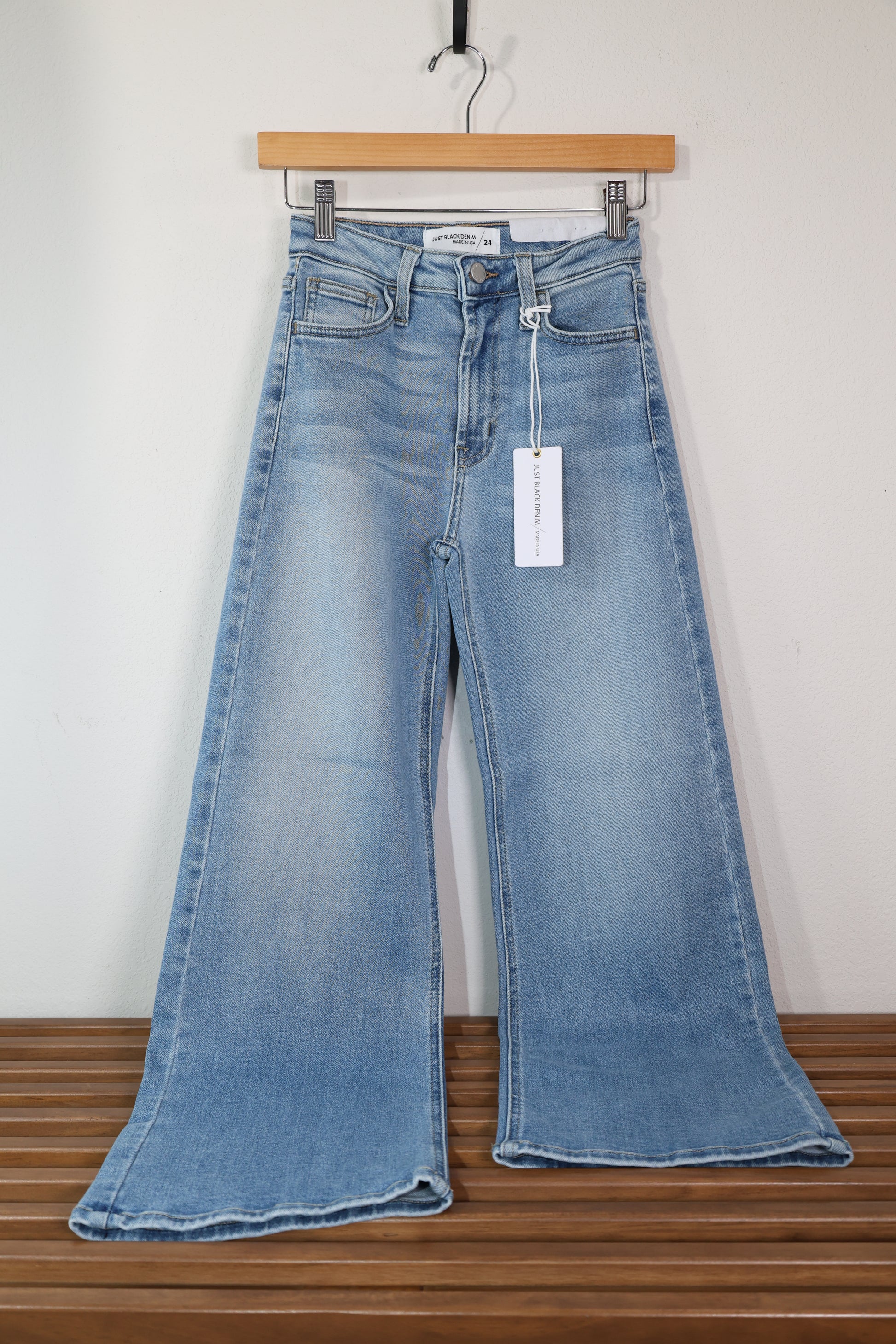 A casual pair of jeans shown displayed on a wooden hanger, the jeans are a light wash blue jean with a slim wide leg and finished hem, the wash is perfect for spring 